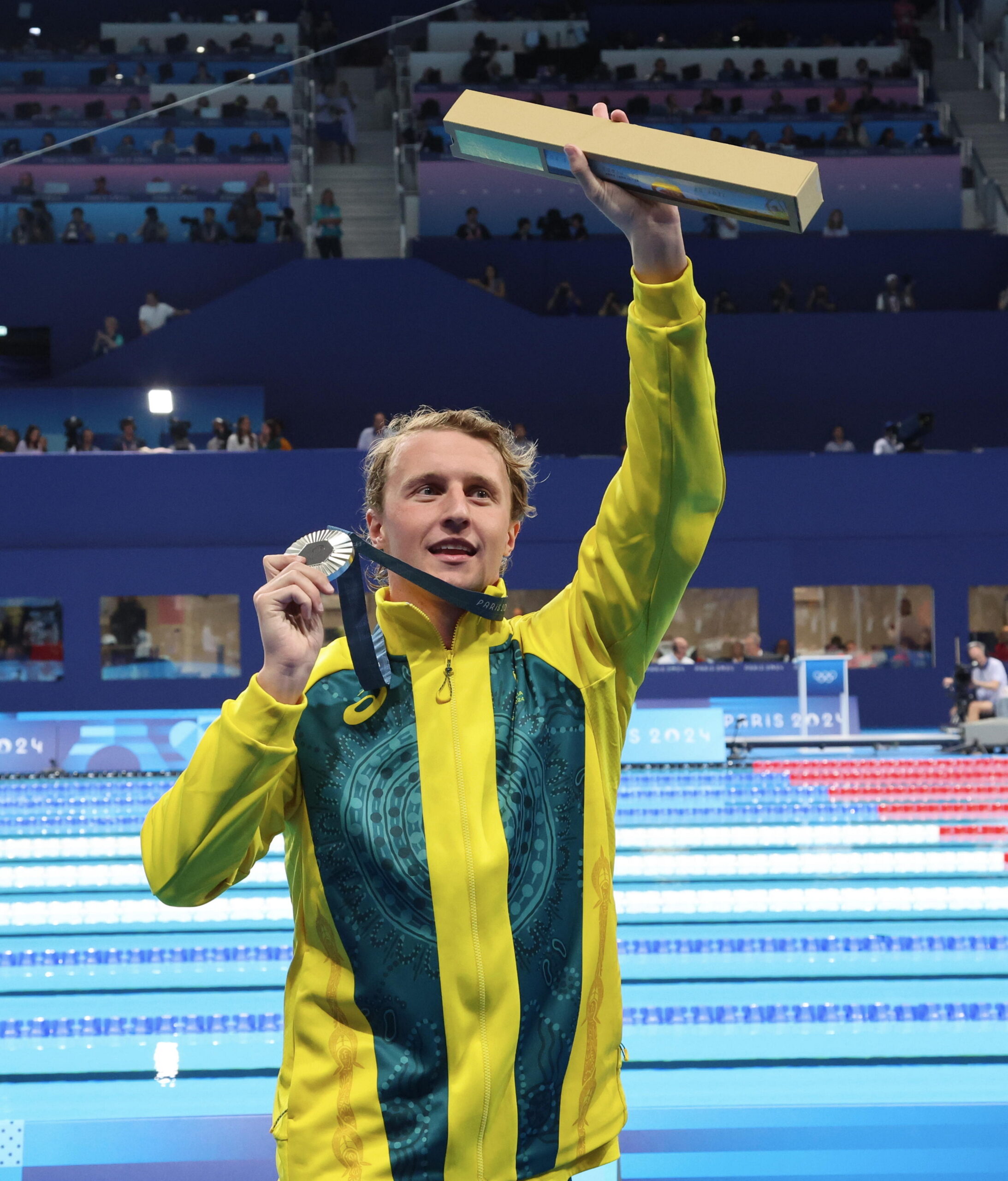 Man in yellow jacket holds silver medal and trophy at swimming pool, celebrating at an indoor sports event.