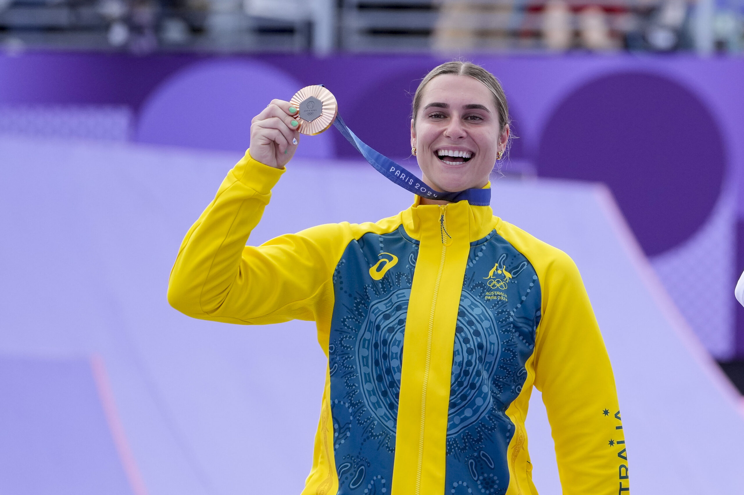 Athlete in yellow Australian jacket smiling, holds a Paris 2024 medal in front of a purple background.