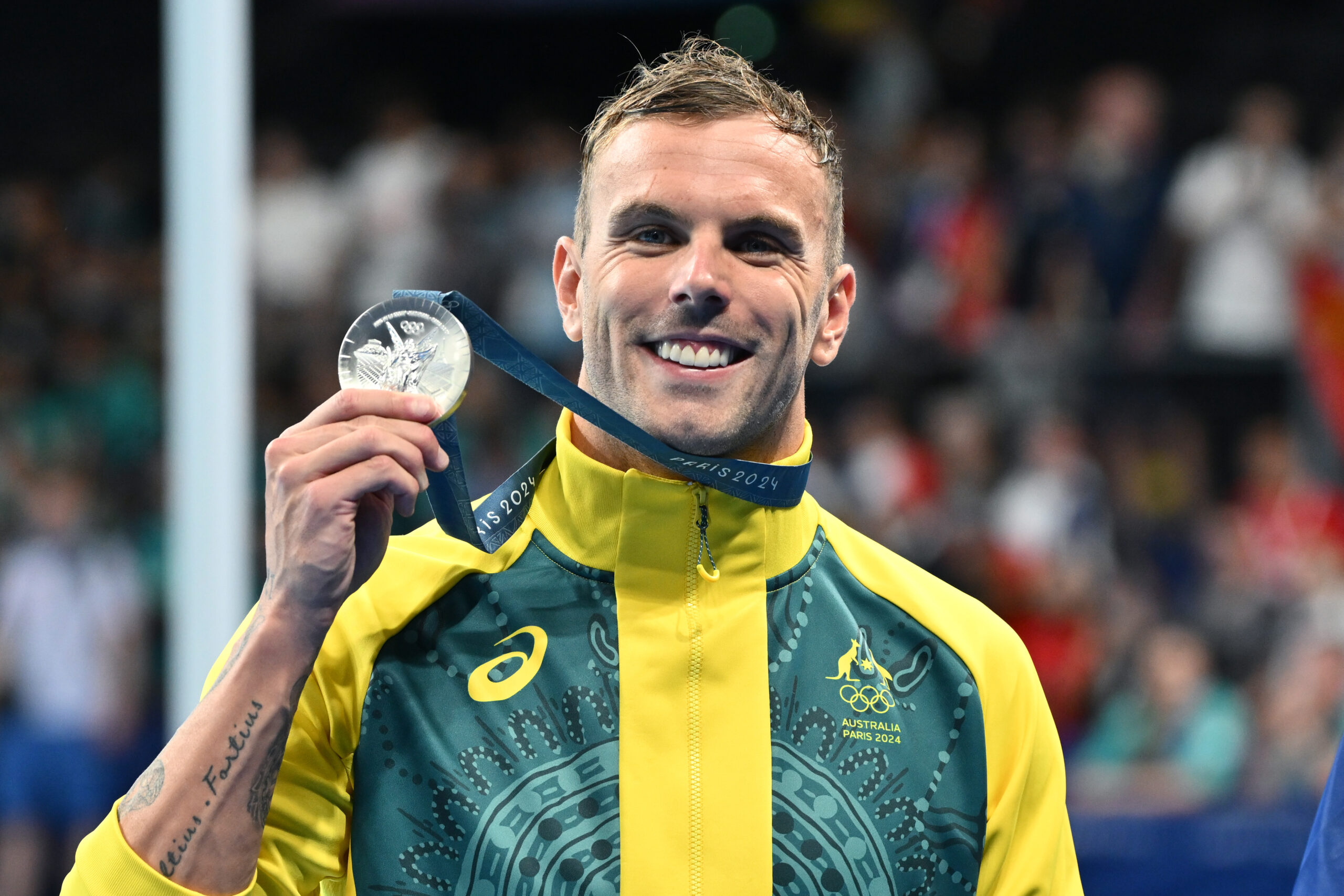 Olympian in Australian tracksuit holding a silver medal with "Paris 2024" lanyard, smiling at the camera.