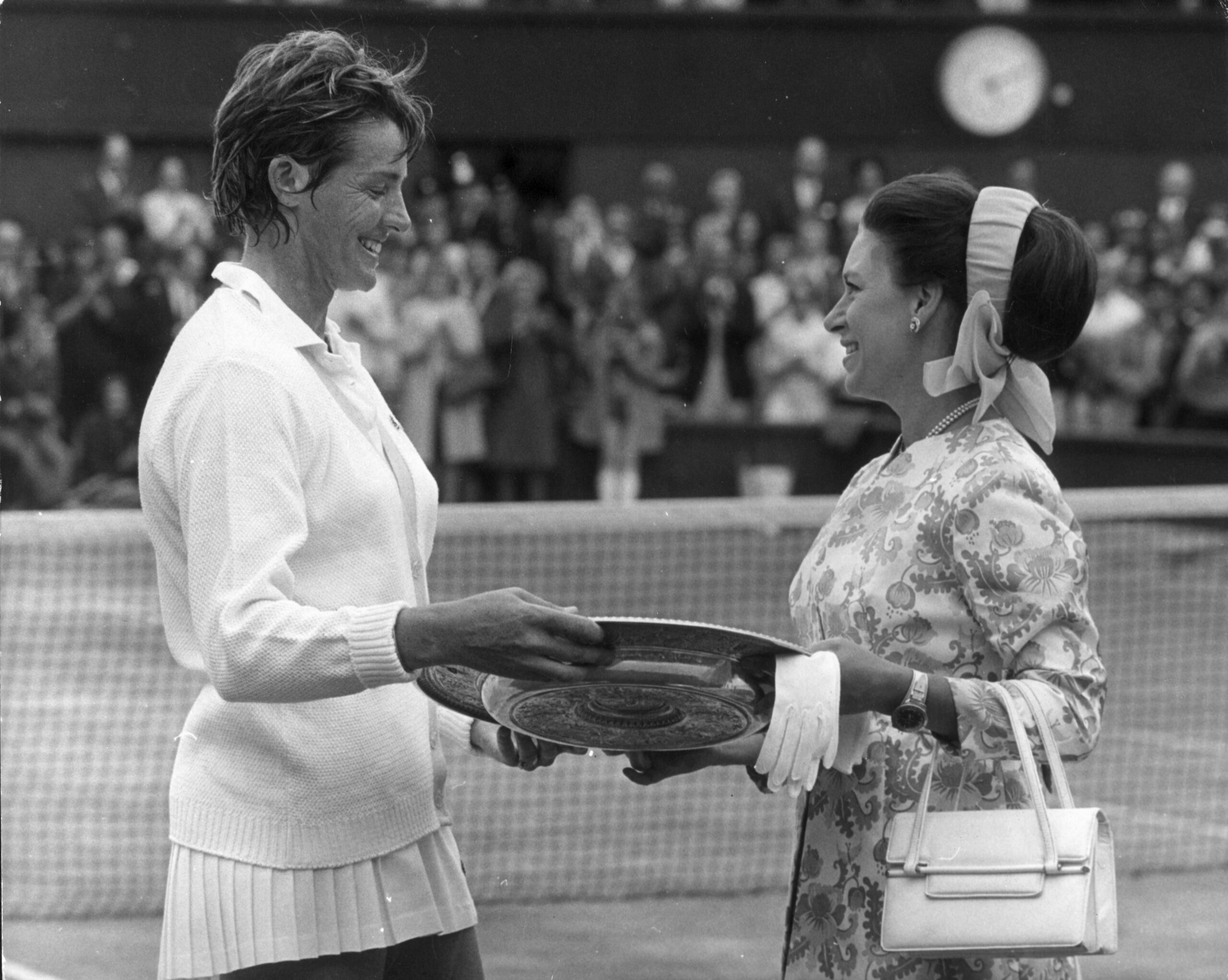 Tennis player accepts trophy from a woman at a court ceremony, with a crowd in the background applauding.