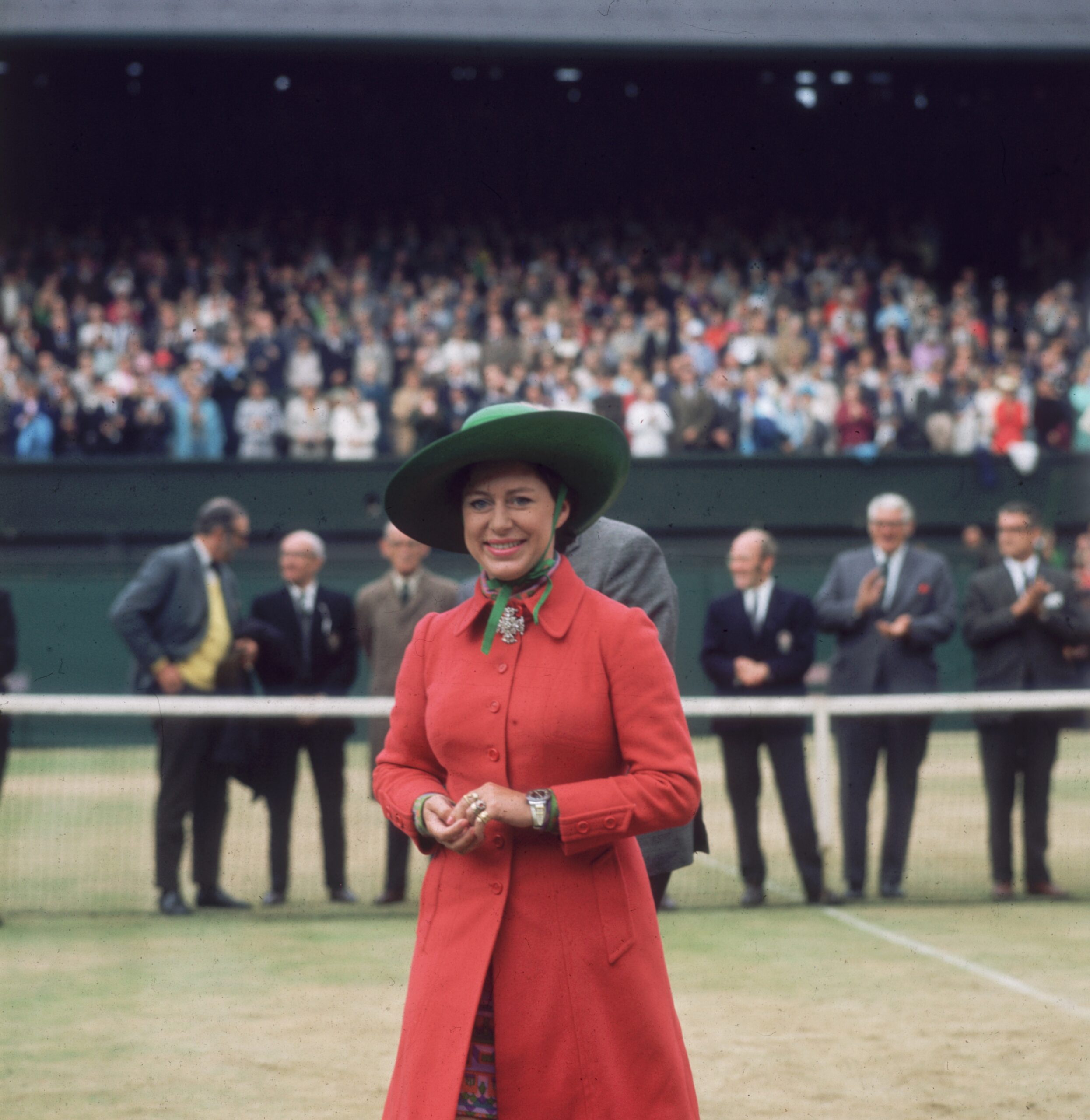 A woman in a red coat and green hat stands on a tennis court with a crowd in the background at Wimbledon.