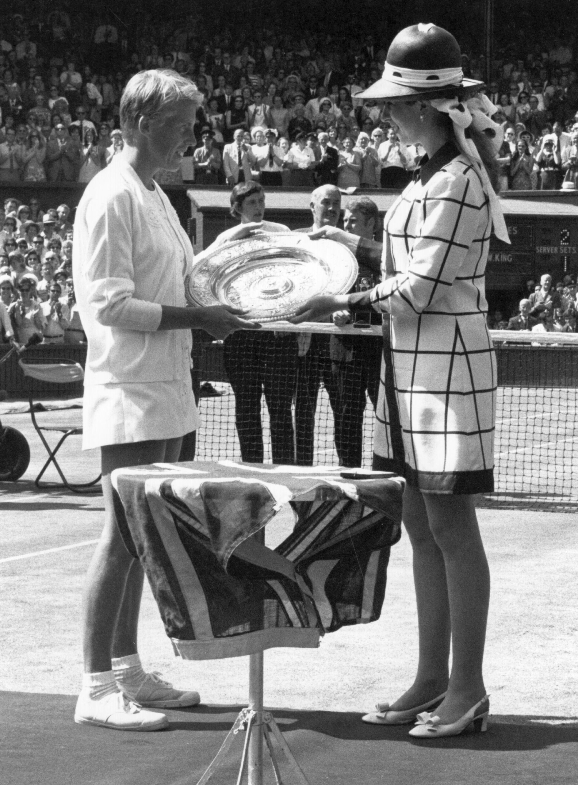 Tennis player receives trophy from a woman in a hat at a crowded tennis court trophy ceremony.