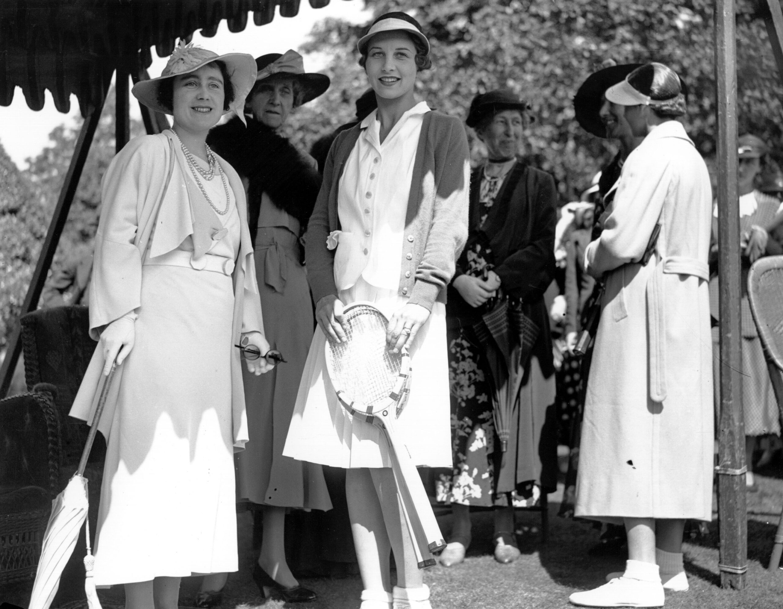 Women in 1930s attire with tennis rackets and parasol, standing outdoors at a social or sporting event.