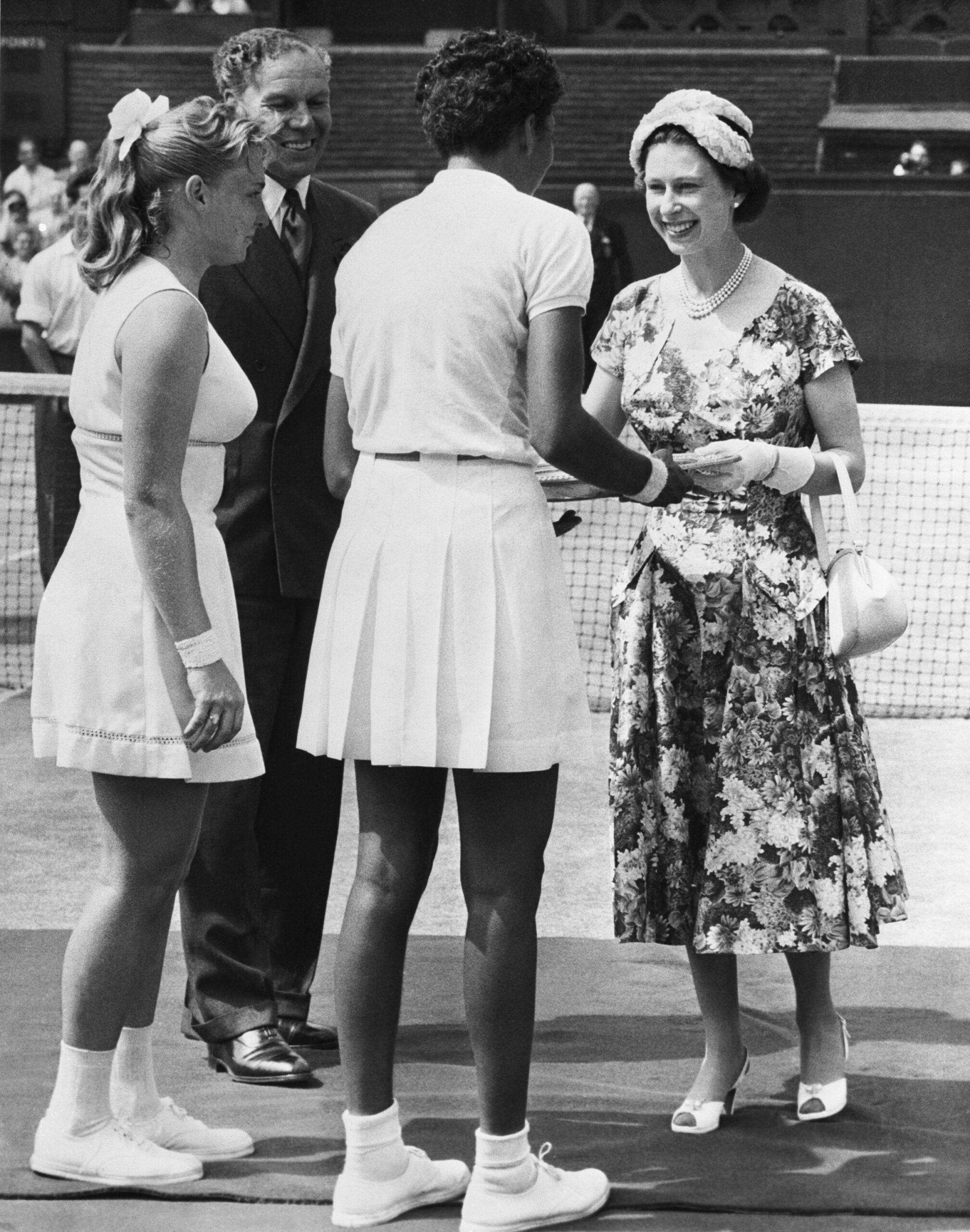 A woman in a floral dress shakes hands with tennis players at a grass court event, with a man smiling nearby.