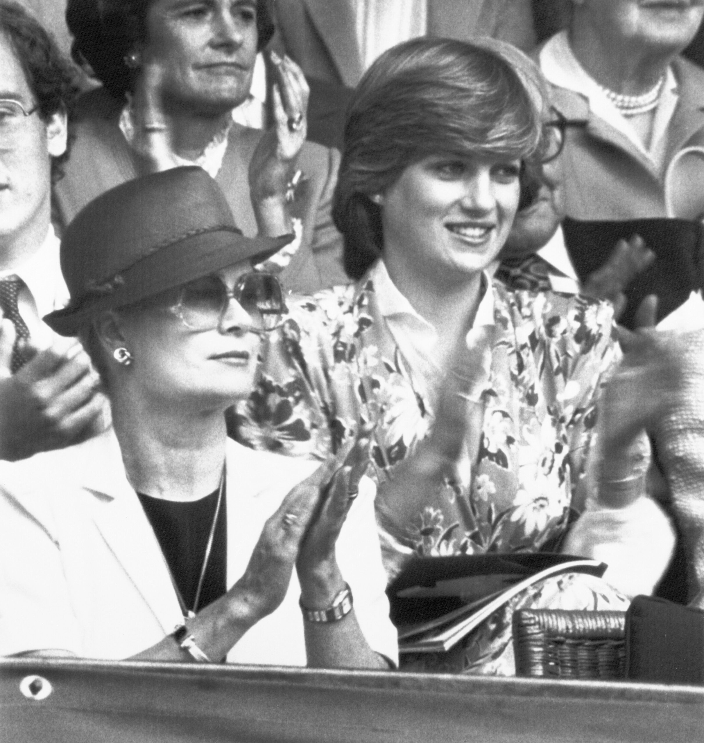 Two women clapping in the audience, one wearing sunglasses and a hat, the other in a floral dress, surrounded by others.