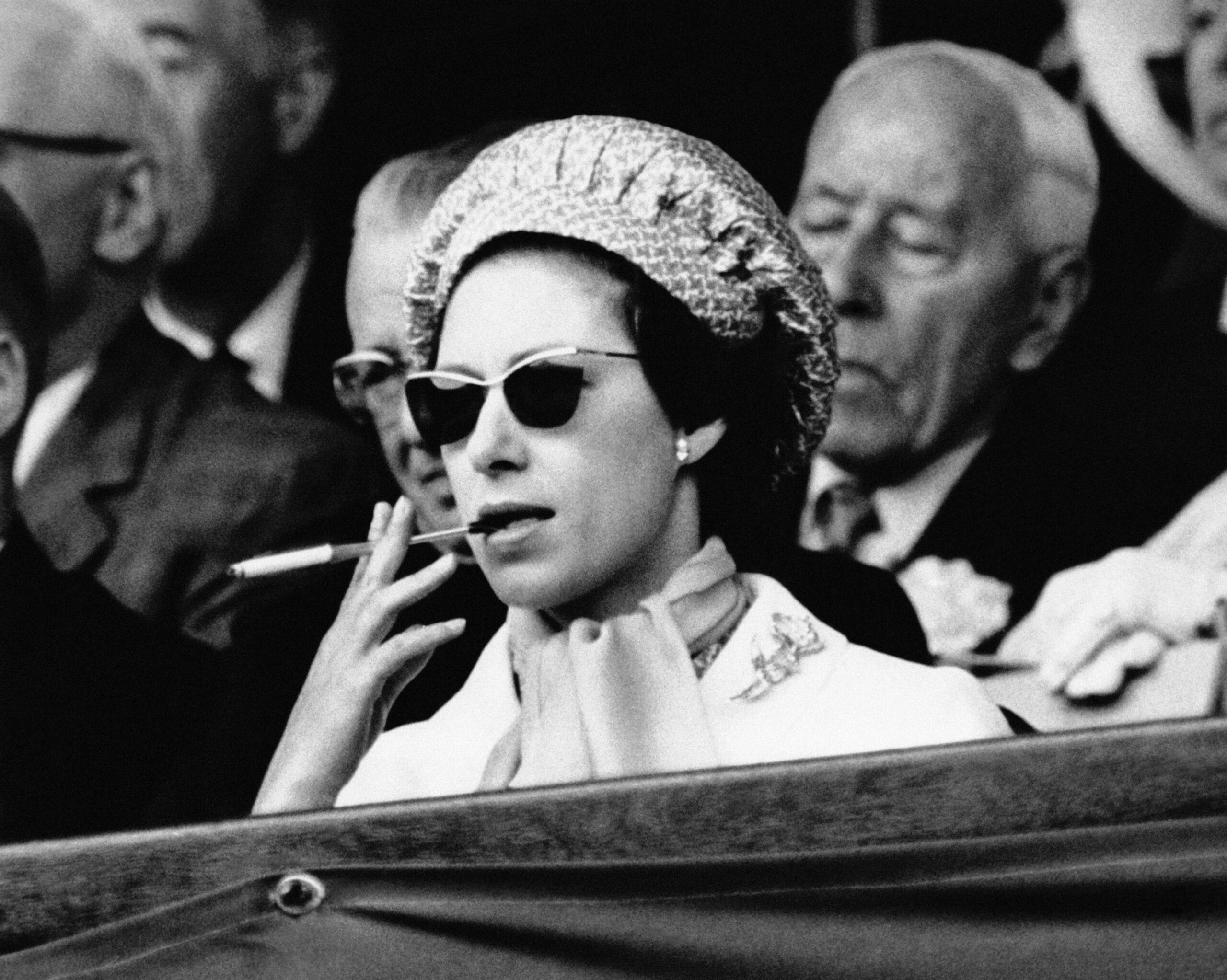 A woman in sunglasses and a hat smokes while sitting in a crowd at an event.