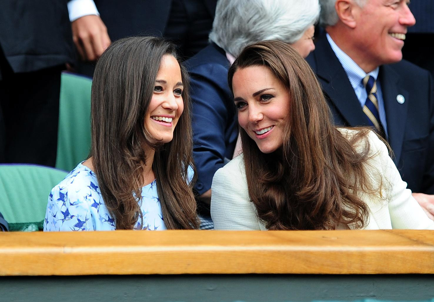 Two women with long brown hair smiling at a sports event, seated in a row with people in the background.