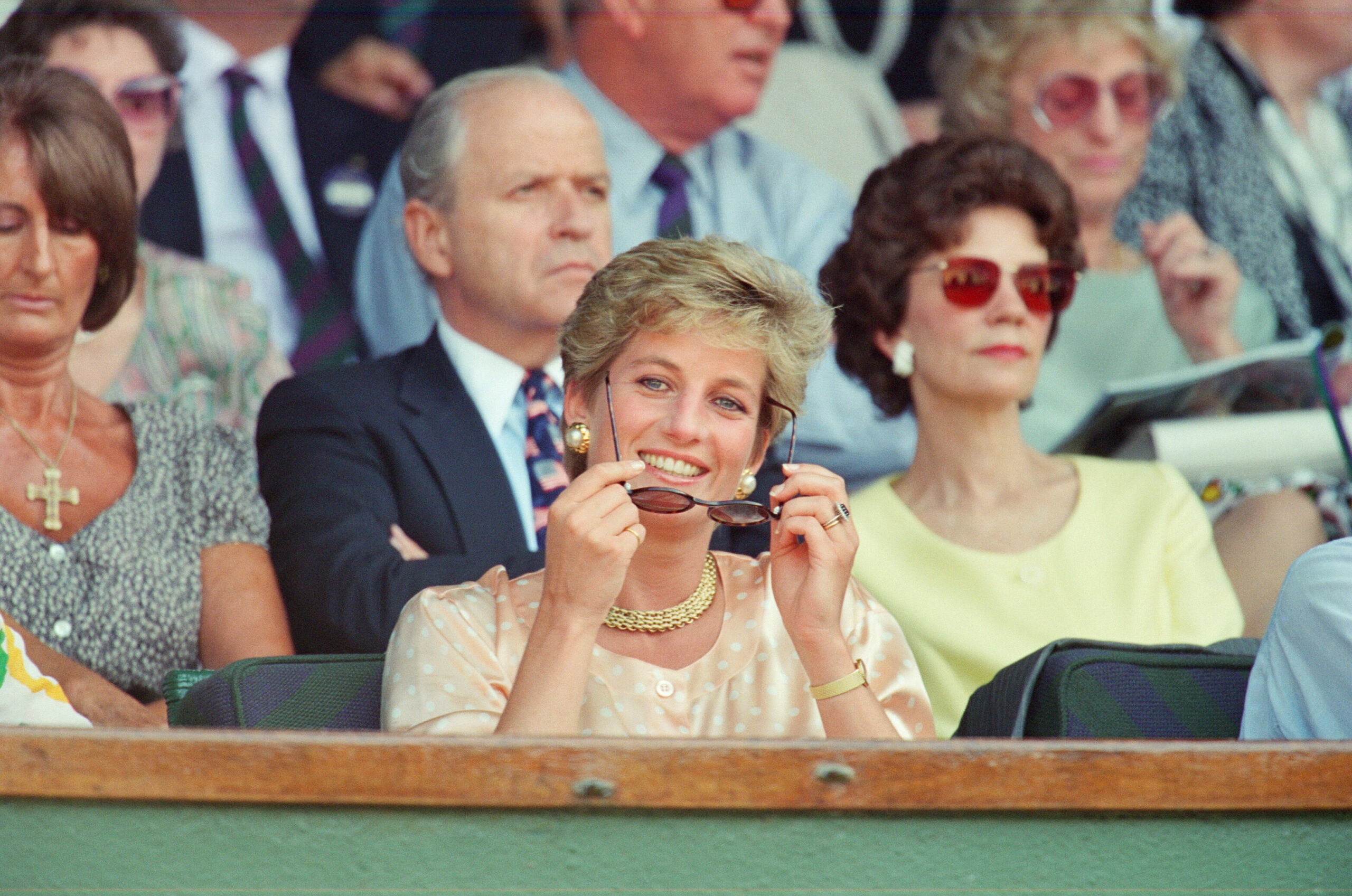 A woman in a light outfit smiling and holding sunglasses, sitting among a crowd at an outdoor event.
