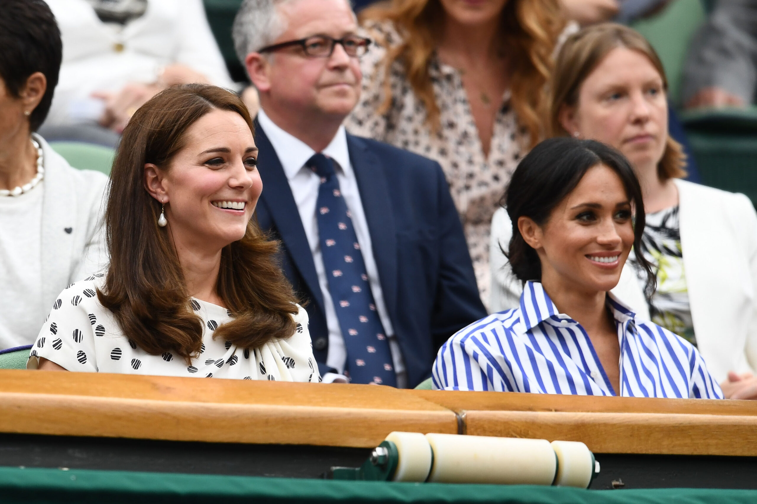 Women in a stadium seating area, one in a white patterned dress and the other in a blue striped shirt, smiling at an event.