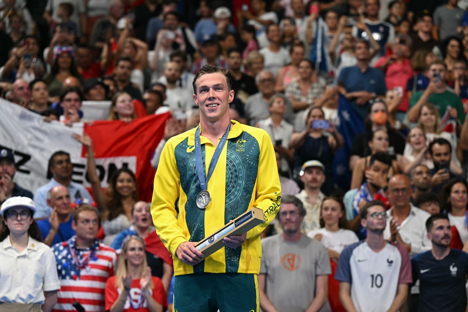 Australian athlete wearing a medal and holding a trophy, stands smiling, with applauding crowd in background.