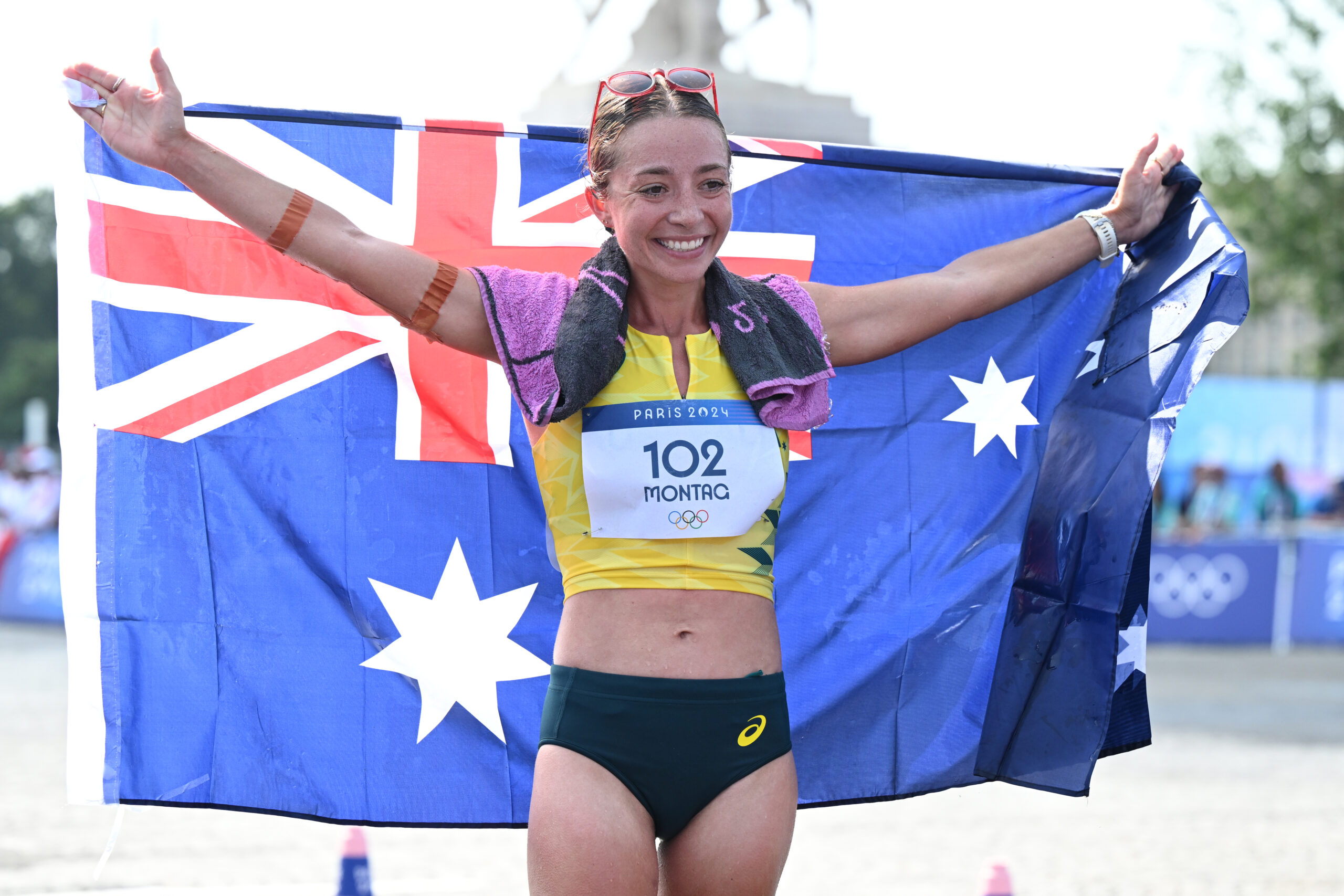 Runner holding an Australian flag, smiling, wearing a yellow and green kit with number 102, labeled "Montag," at Paris 2024.
