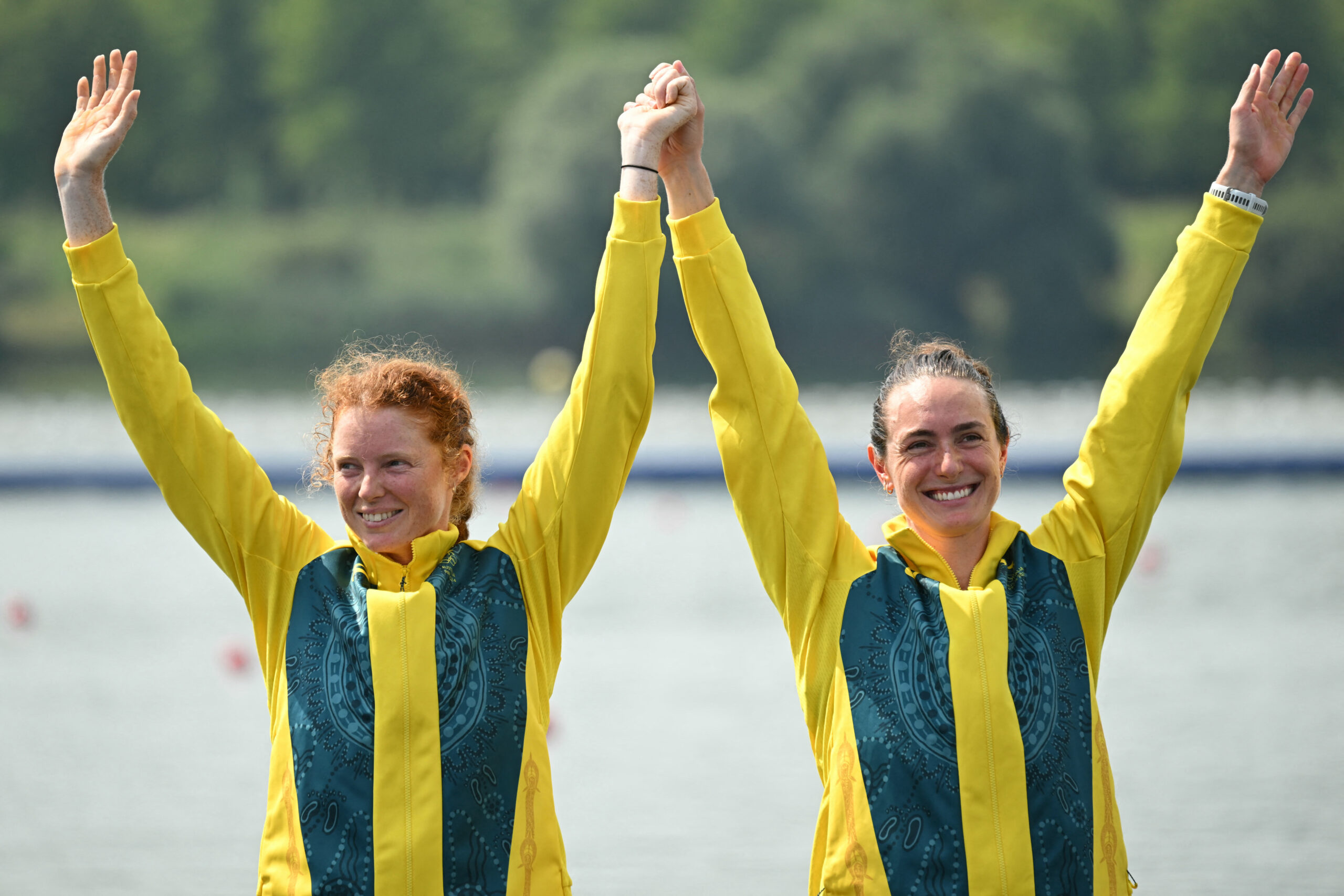 Two female athletes in yellow jackets smile and raise their hands in victory by a water body.