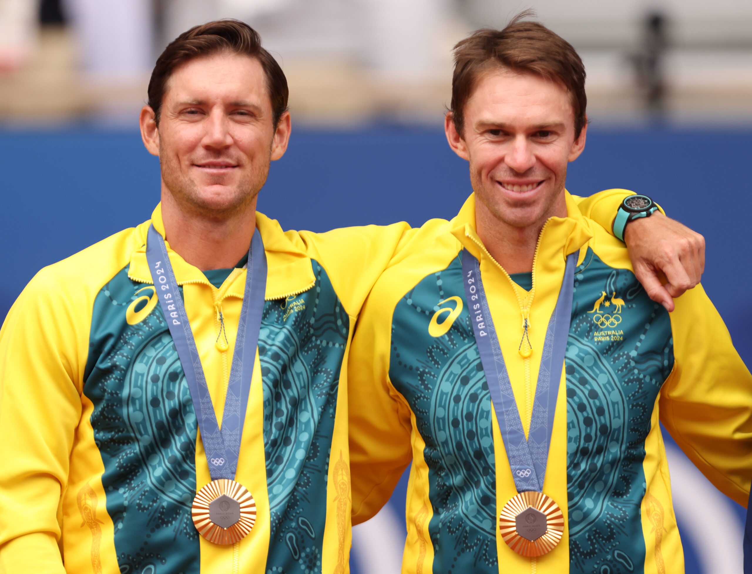 Two athletes in yellow-green outfits with bronze medals at an Olympic event, smiling with arms around each other.