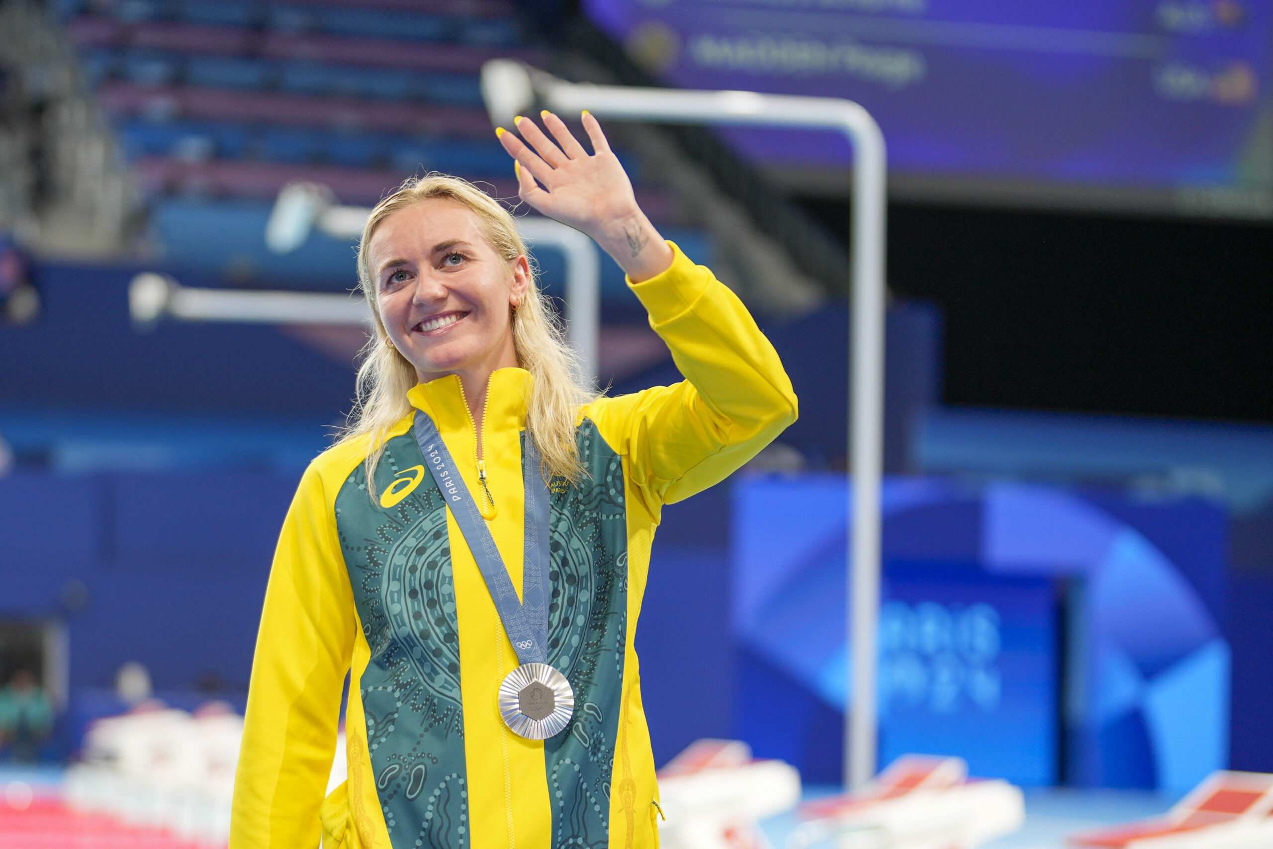 Athlete in yellow and green jacket with a silver medal waves, smiling, indoors at a sporting event.