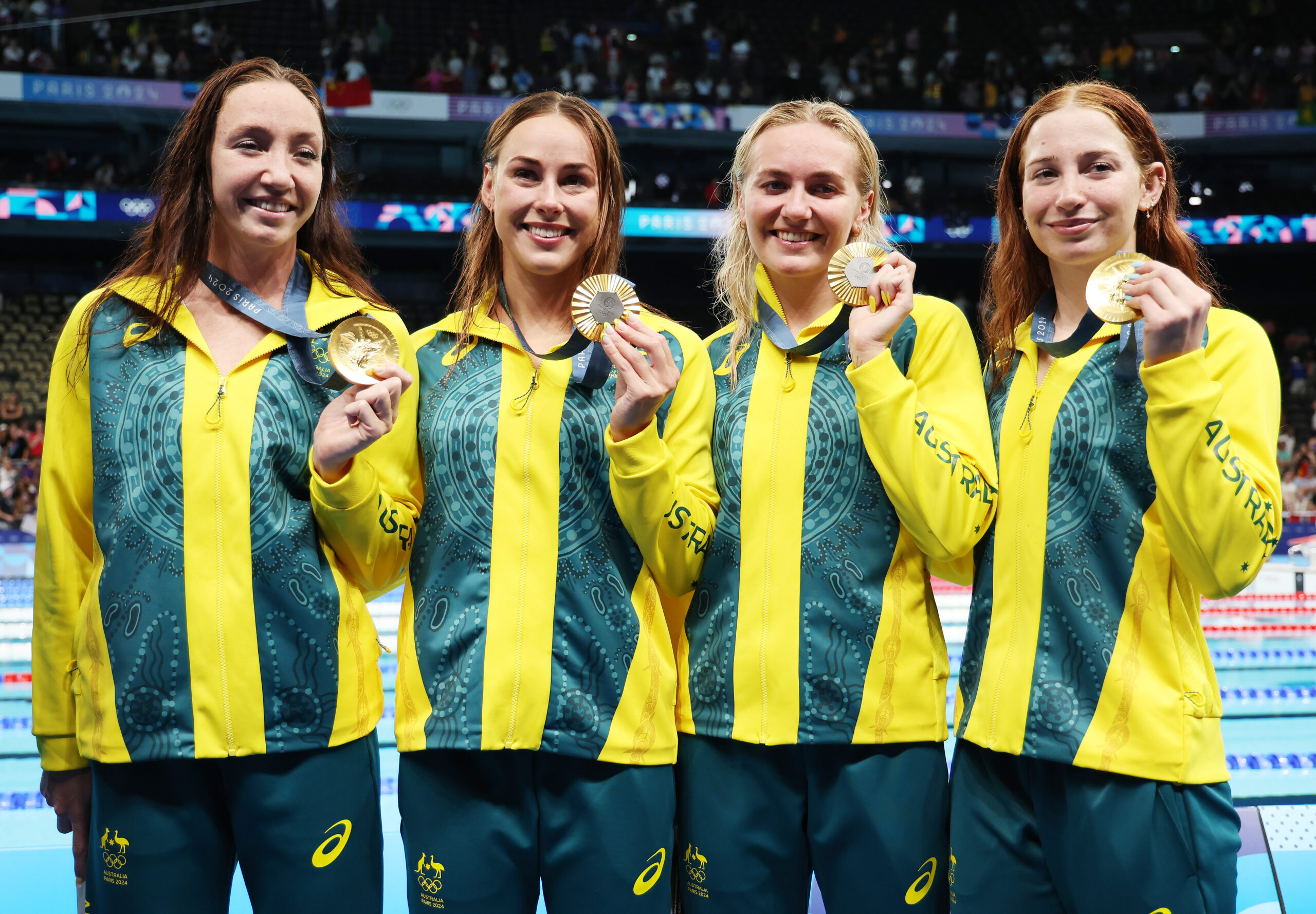 Four female athletes in Australian uniforms holding gold medals at a swimming event.