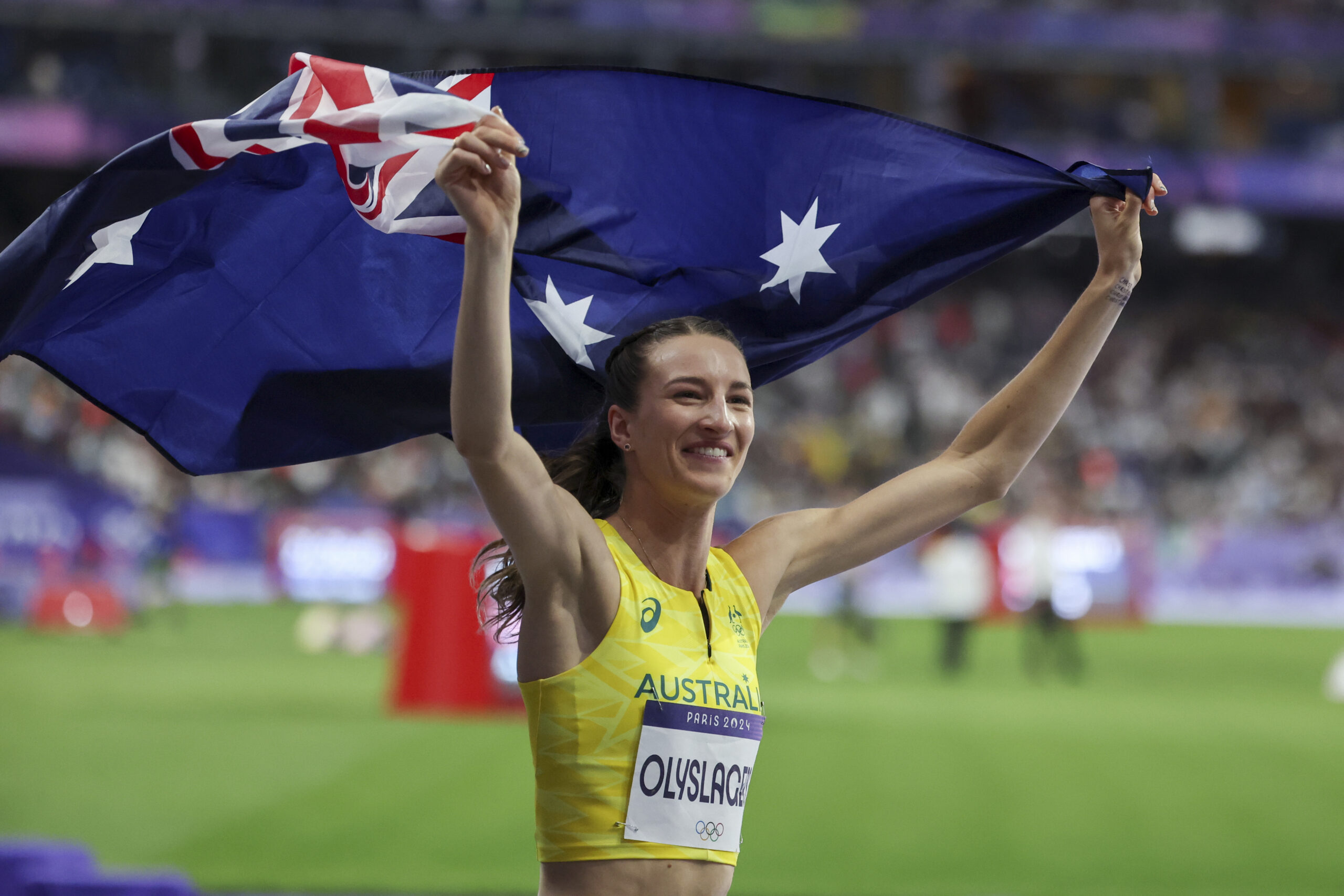 Athlete celebrates with Australian flag in a stadium, wearing a yellow jersey with "Olyslagers" written on it.