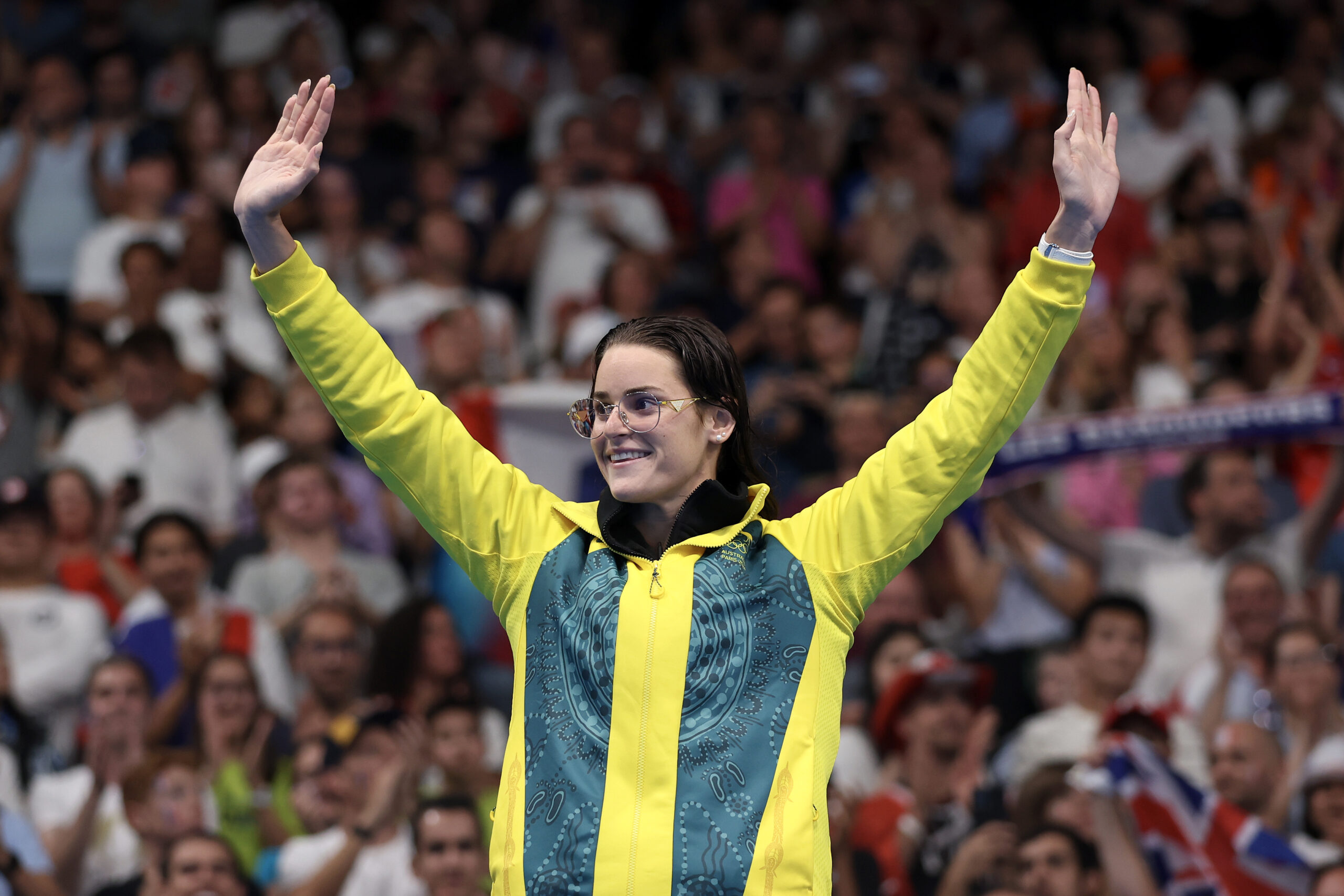 A person in a vibrant jacket raises their arms in triumph, smiling at a cheering crowd.