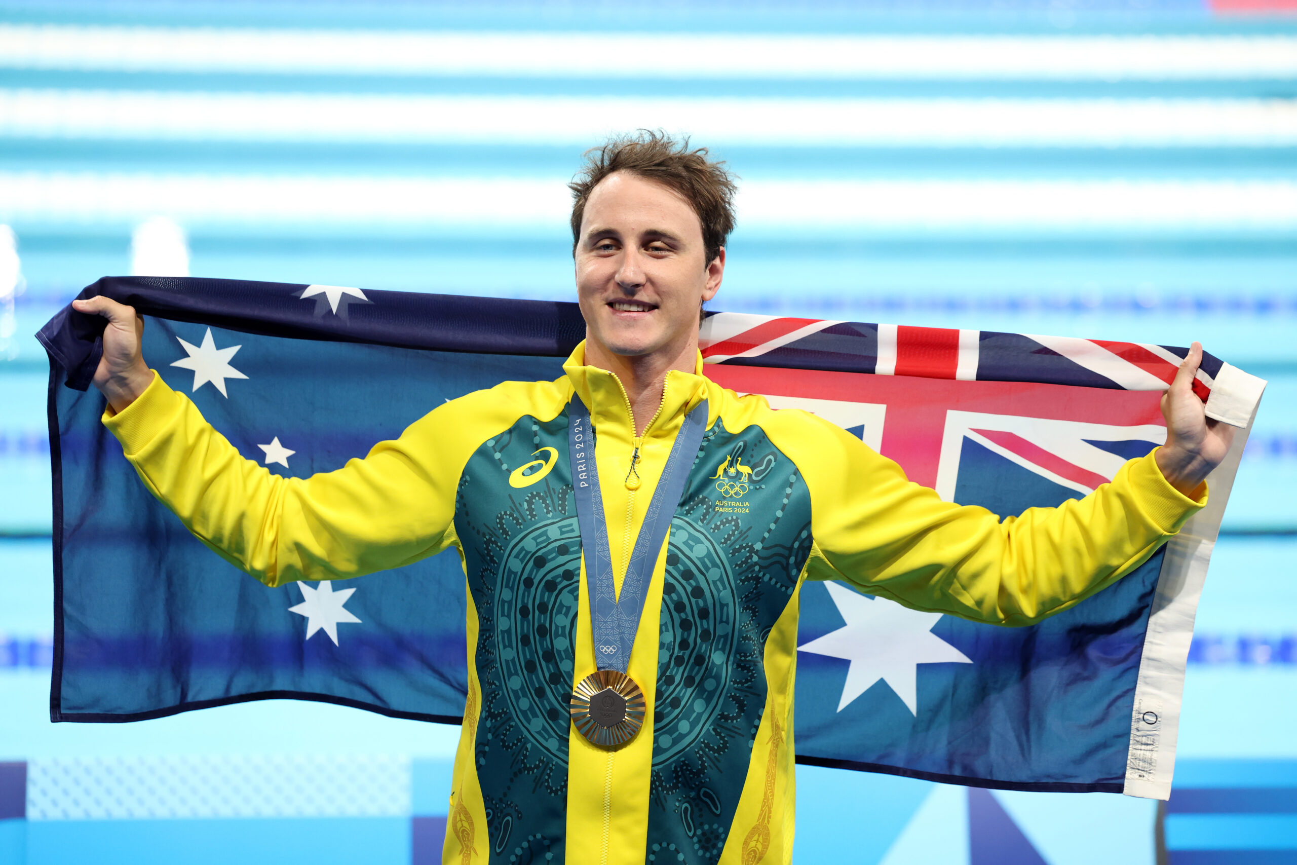 Australian athlete holding a medal and flag, wearing a green and yellow tracksuit, smiling by a pool.
