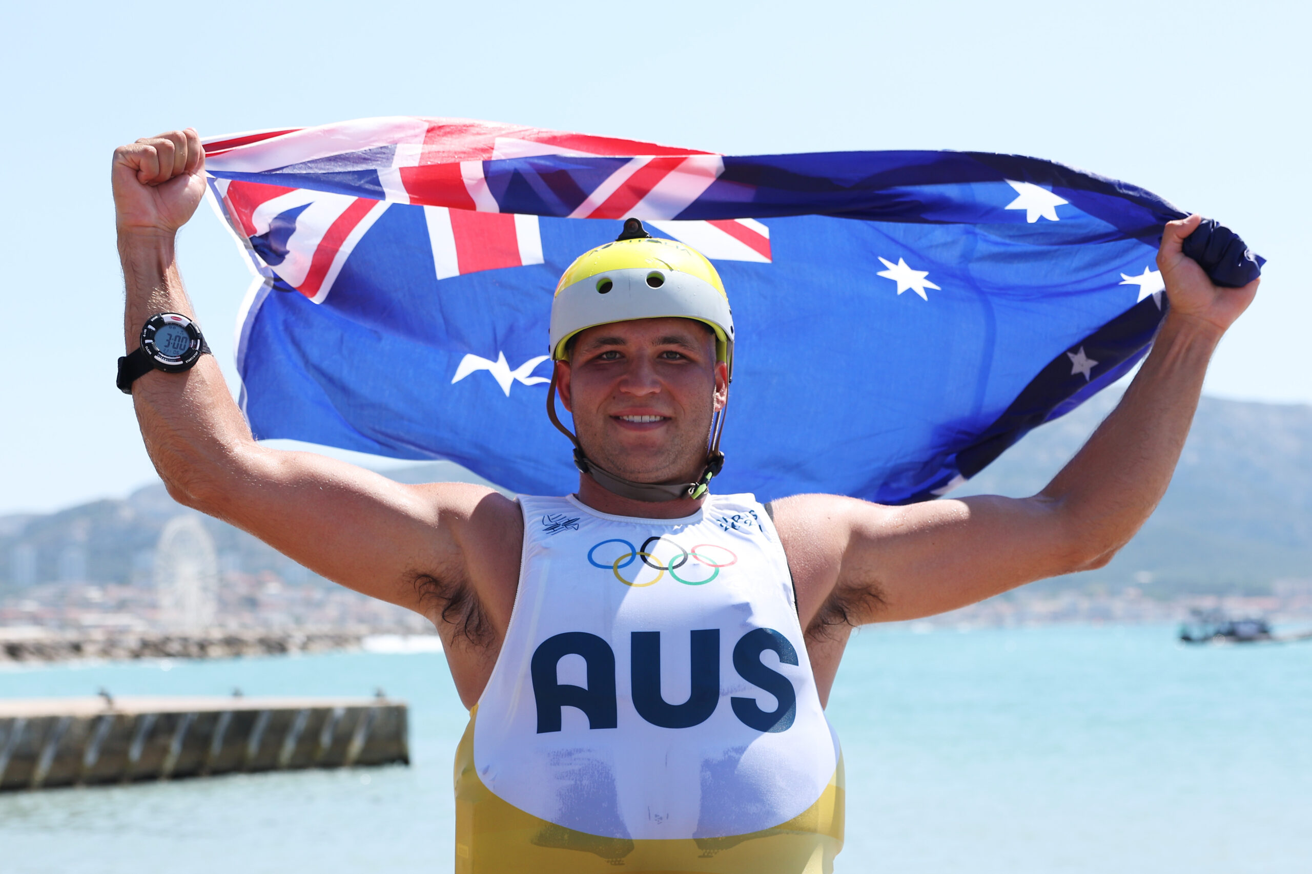 Australian athlete holding flag with Olympic rings on jersey, smiling near water.