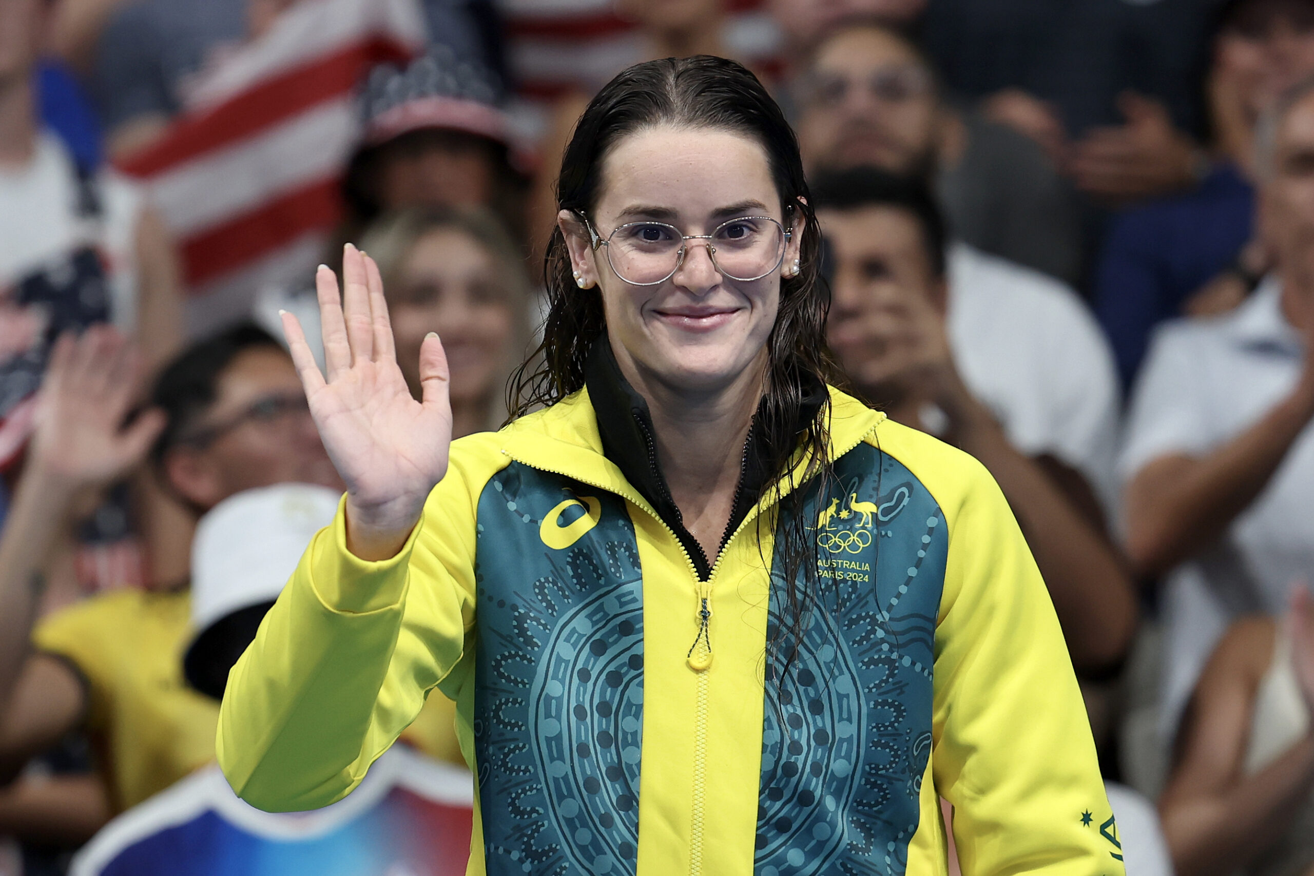 Australian swimmer in glasses, yellow jacket, waves to crowd smiling.