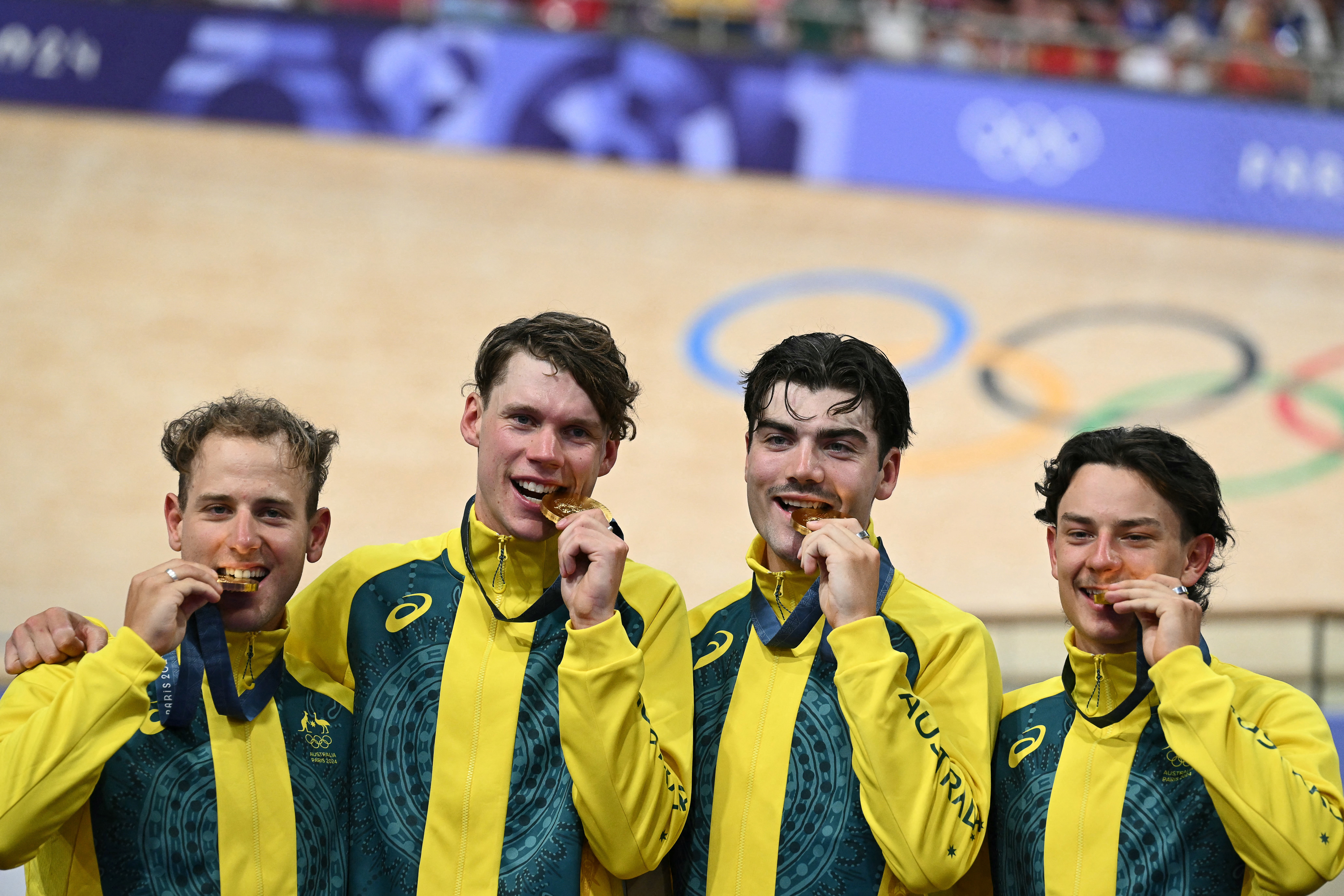 Australian cyclists bite gold medals, celebrating on the Olympic podium in yellow and green jerseys.