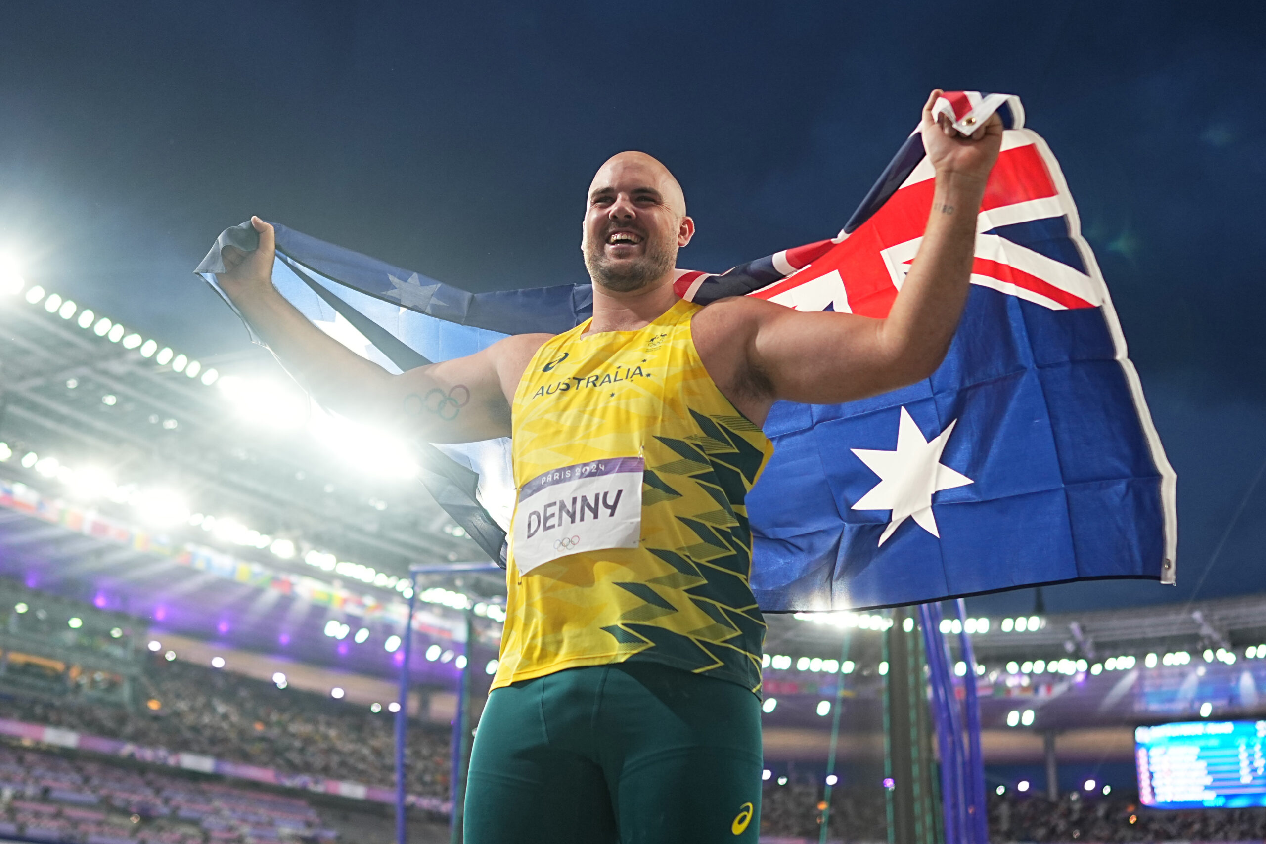 Australian athlete celebrating with flag during 2024 event in stadium, wearing yellow-green uniform labeled "Denny."