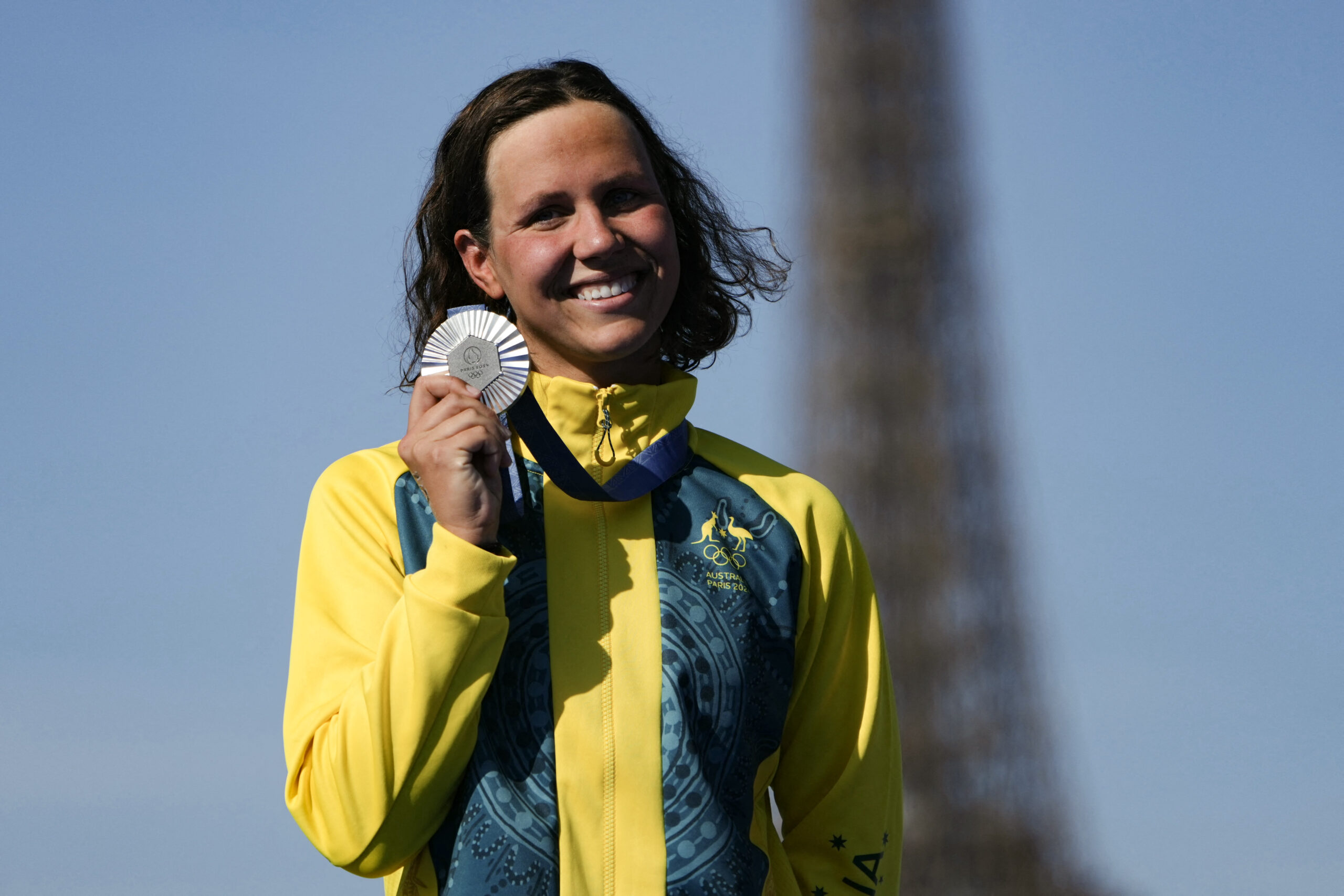 Australian athlete in a yellow jacket smiling and holding a silver medal with the Eiffel Tower blurred in the background.