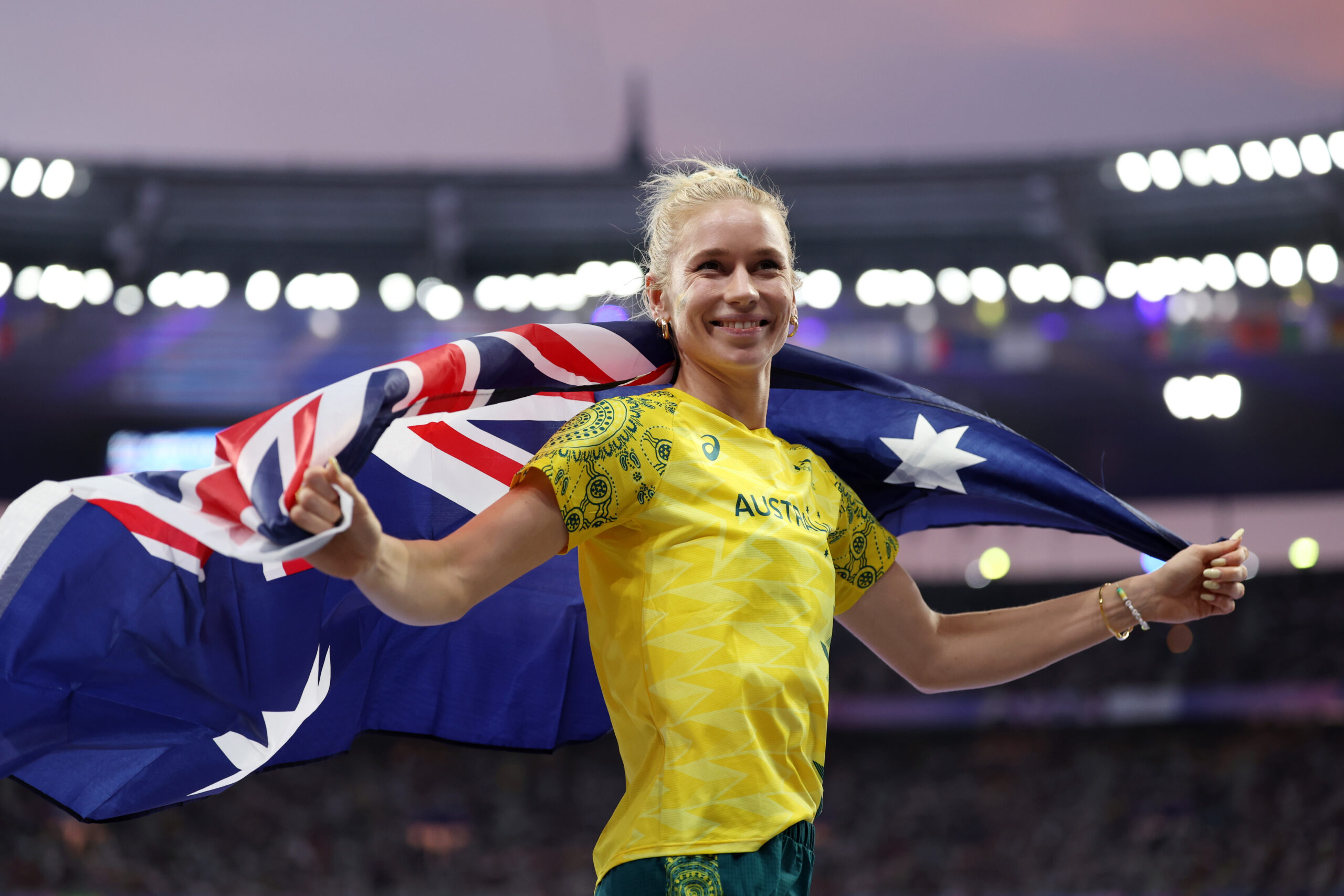 Australian athlete smiling, draped in a flag, celebrates at a stadium event under bright lights and a pink sky.