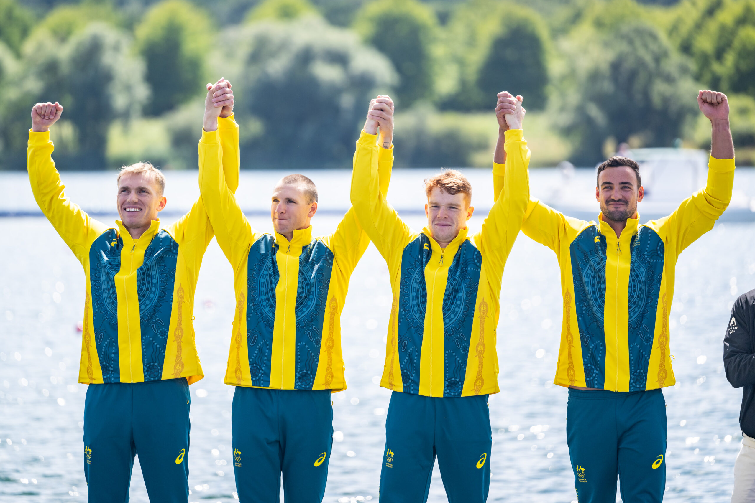 Four athletes in yellow and teal uniforms celebrate by holding hands aloft in front of a lake.