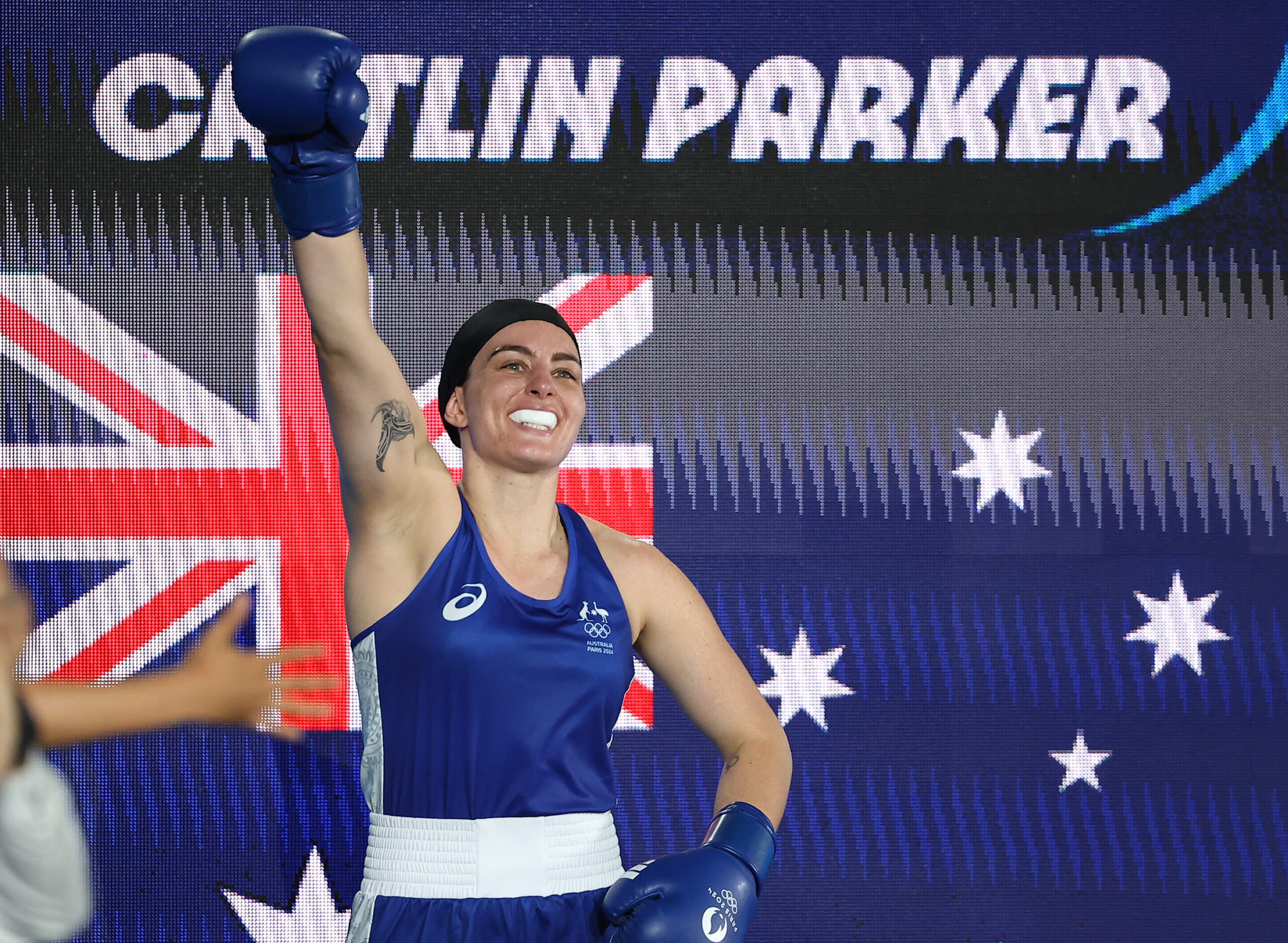 Boxer in blue gear celebrates with raised arm, smiling, against an Australian flag background.